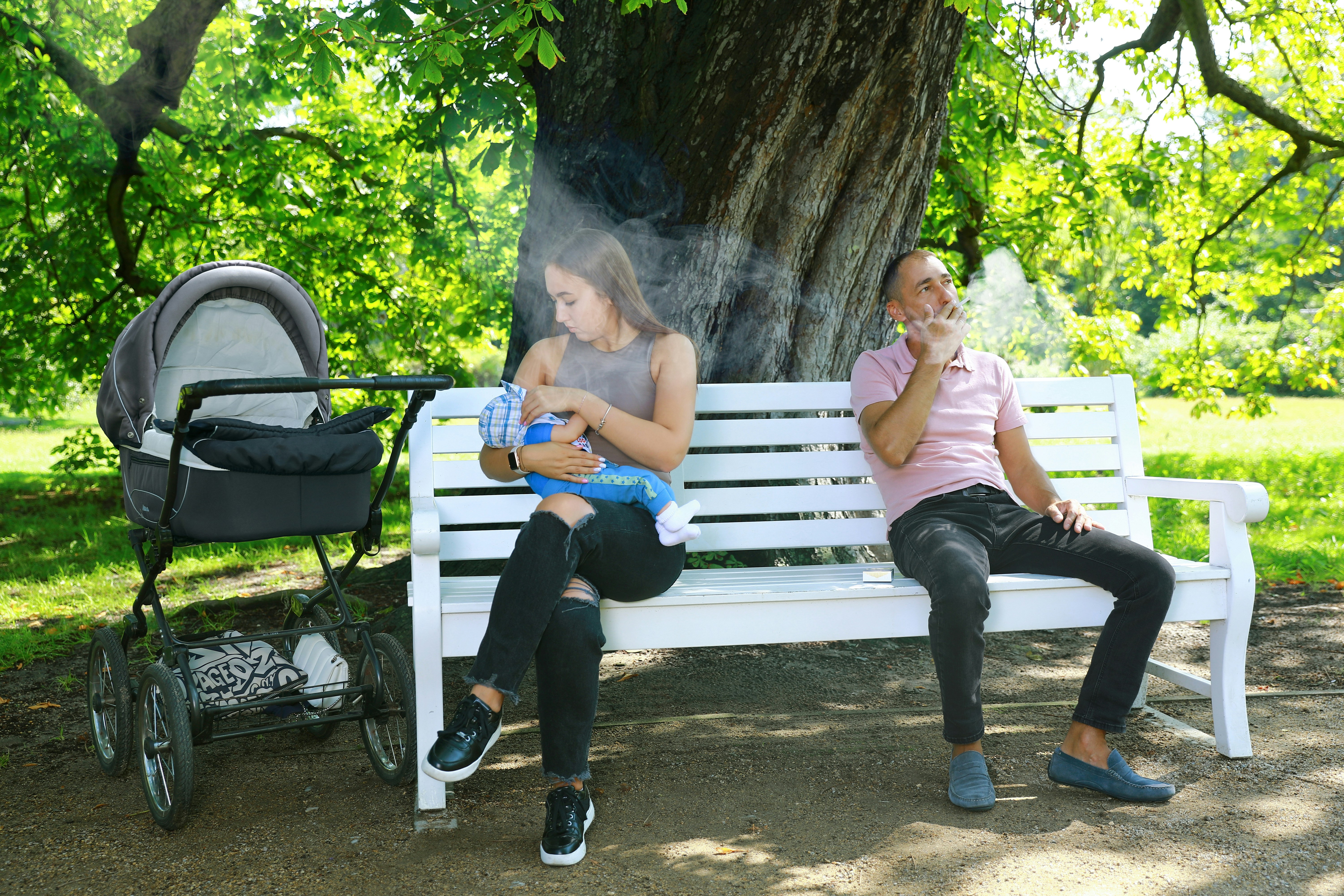 公園長椅上，一名男子在懷抱嬰兒的年輕女子身旁吸菸，煙霧飄向母子兩人