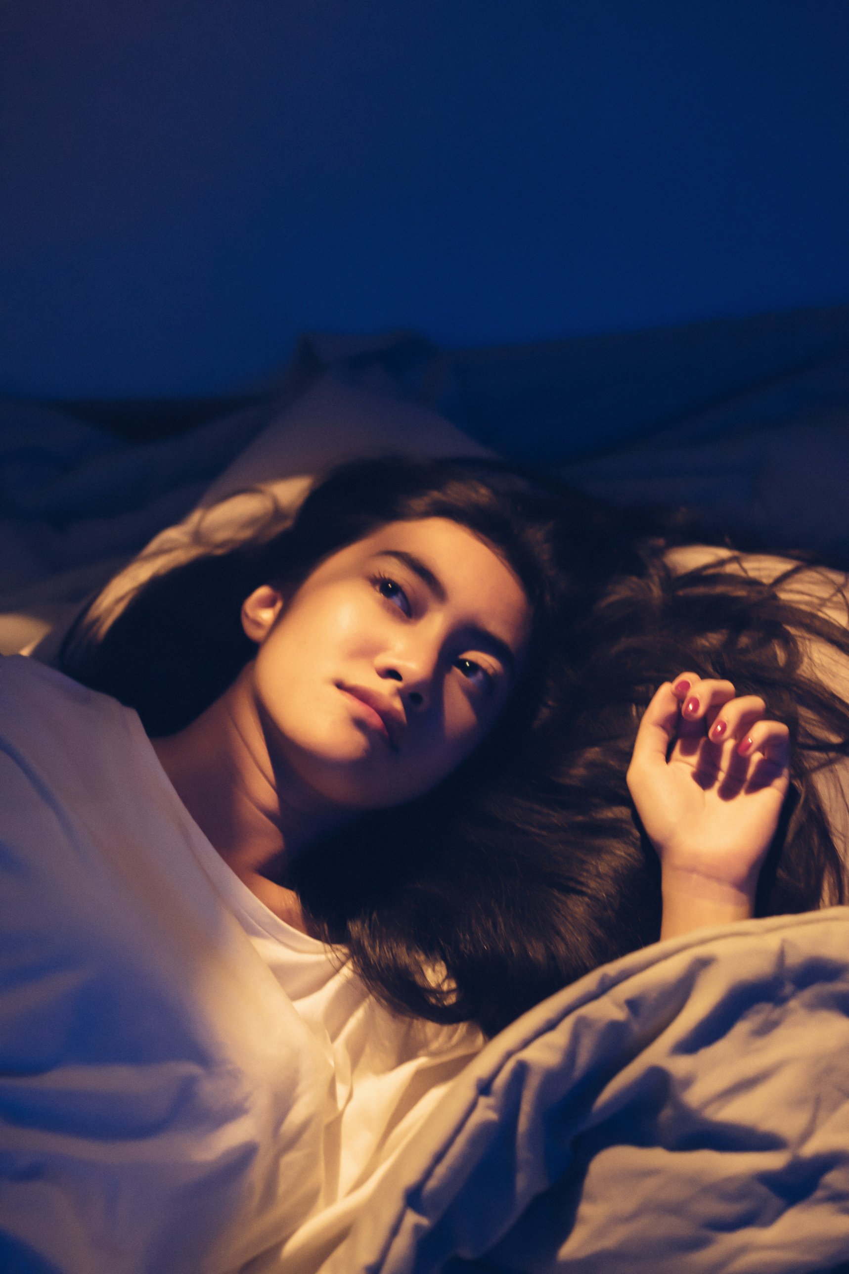 夜裡一名長髮女性躺在床上，蓋著棉被、睜著眼望向上方，臉被暖色燈光照亮