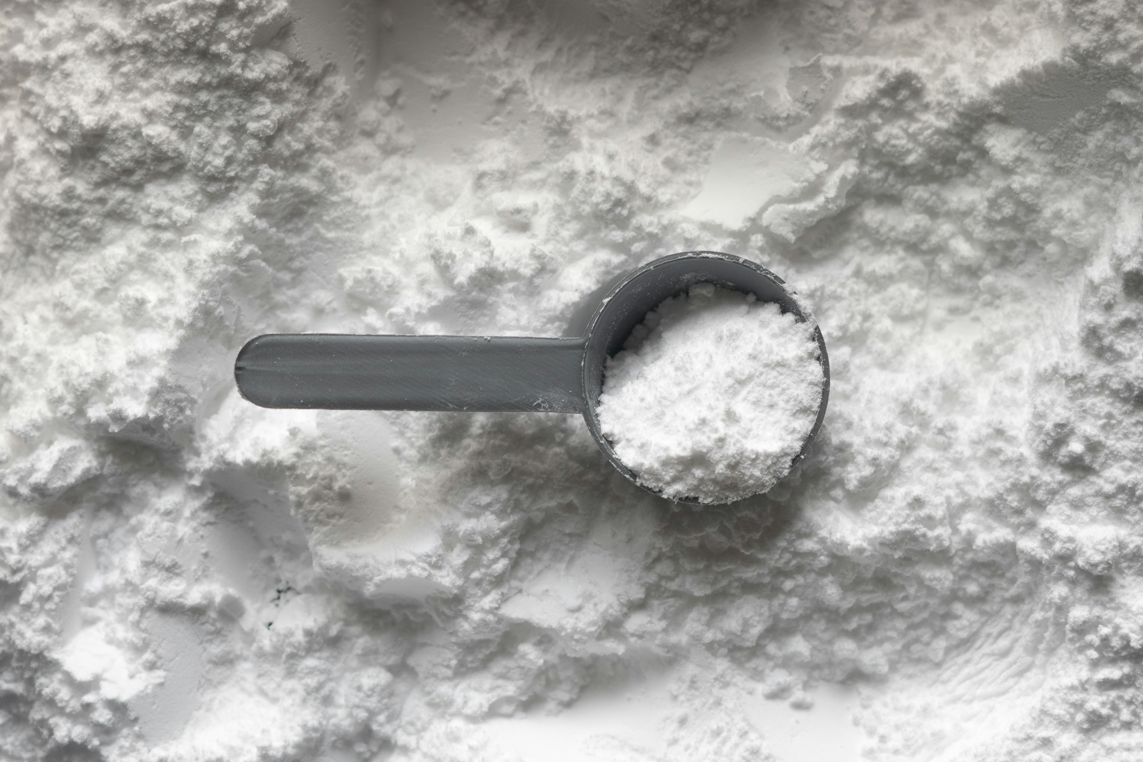 灰色量勺盛著一勺白色肌酸粉末，放在一片灑落的肌酸粉堆上，特寫俯拍