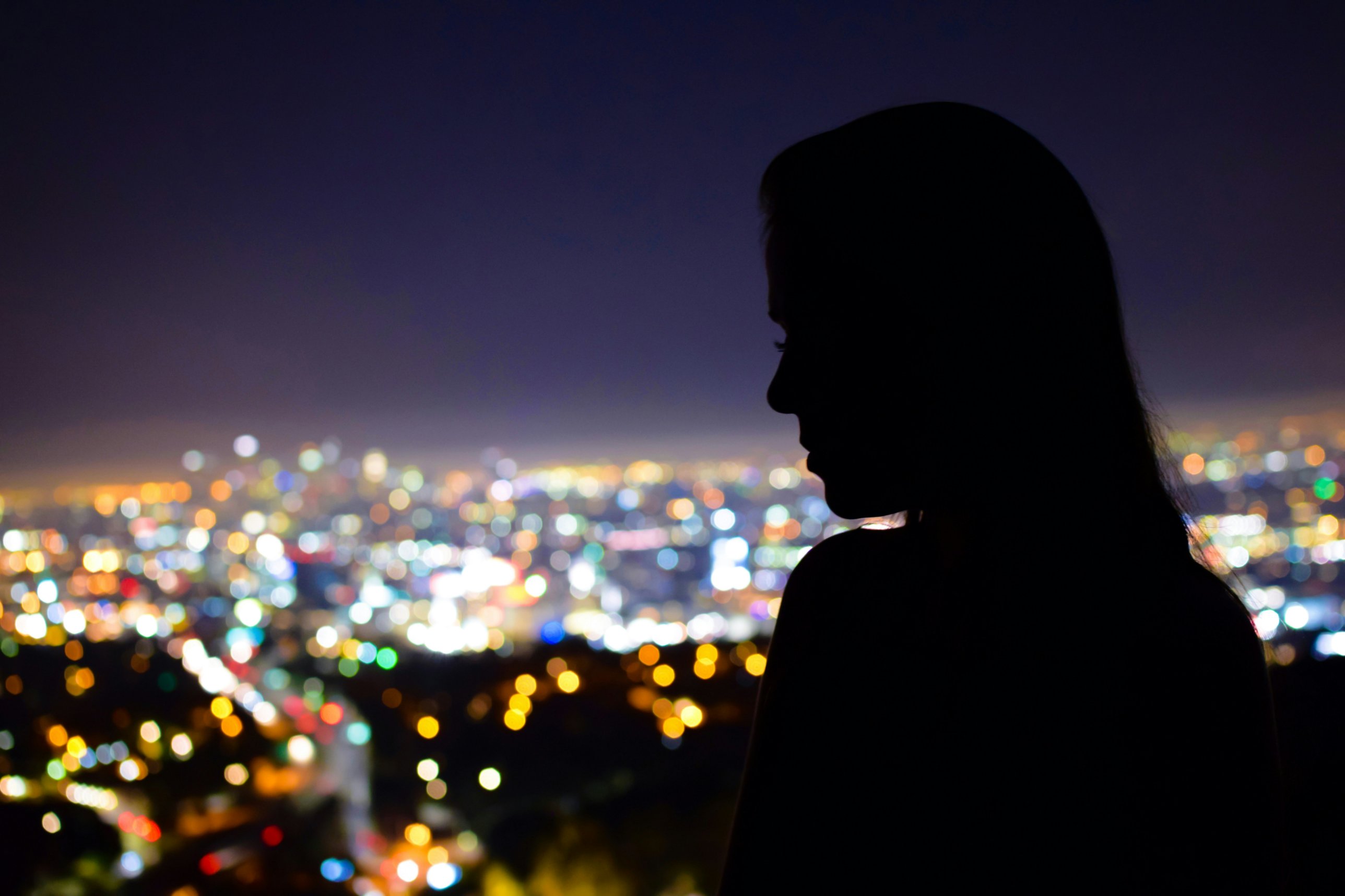 夜晚從高處俯瞰城市燈海，一位長髮女性的側臉剪影位在畫面右側，低頭望著遠方失焦的彩色光點