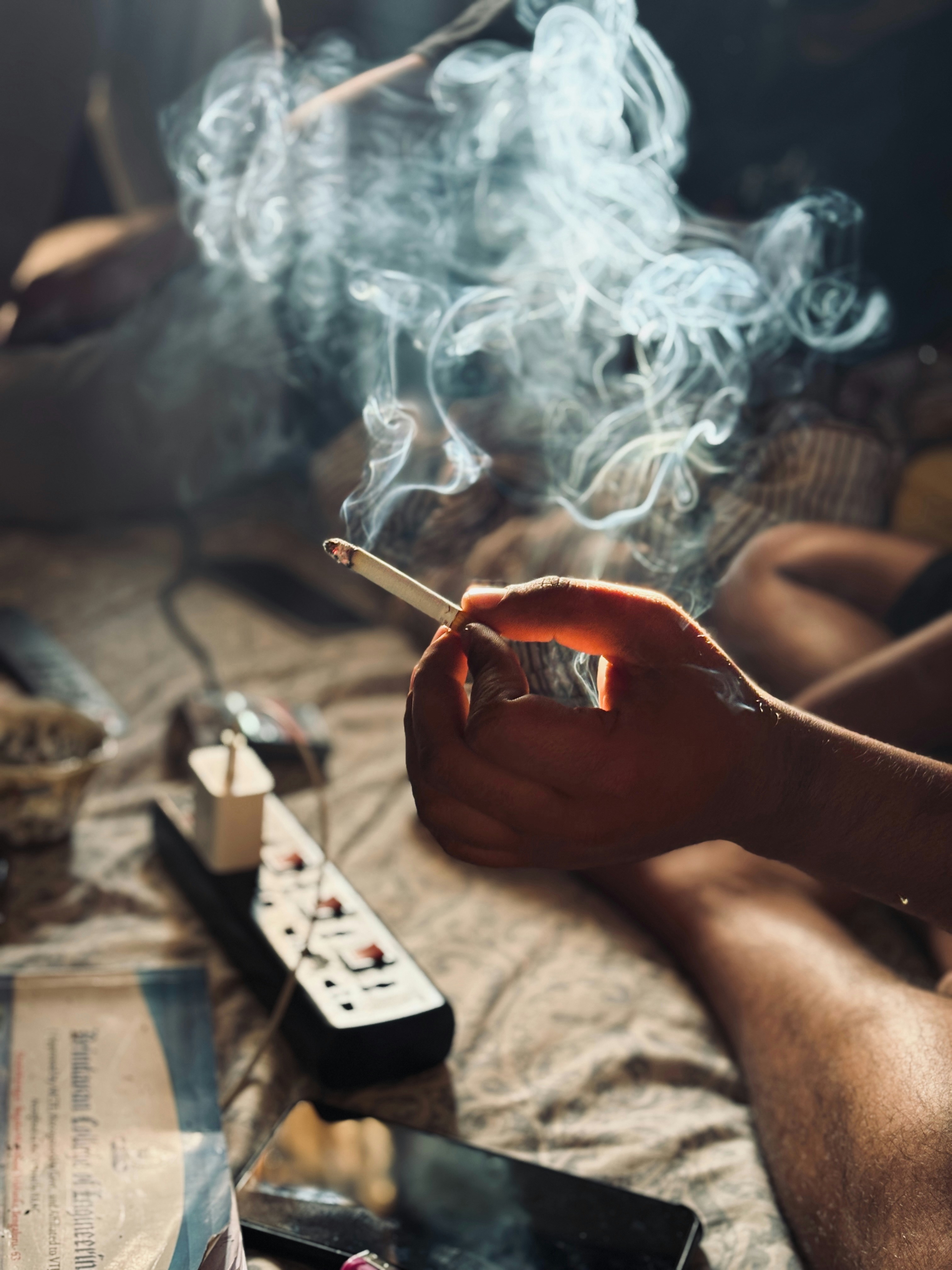 室內昏暗光線中，一隻手拿著點燃的香菸，煙霧在密閉空間裡繚繞擴散，桌上散落著延長線與手機